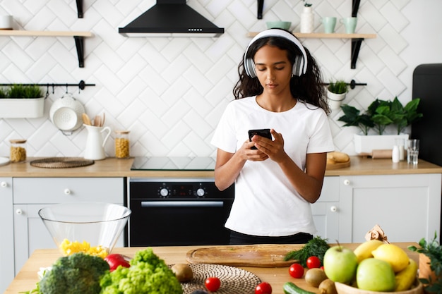 A mulata bonita está segurando o smartphone, em grandes fones de ouvido sem fio, vestindo camiseta branca, perto da mesa com legumes frescos