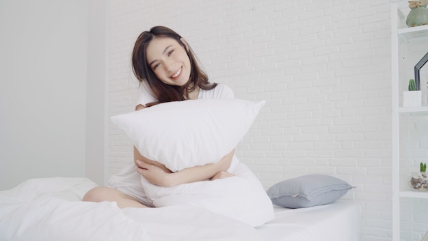 A mulher asiática bonita feliz acorda, sorrindo e esticando seus braços em sua cama no quarto.