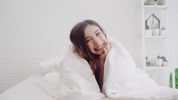 A mulher asiática bonita feliz acorda, sorrindo e esticando seus braços em sua cama no quarto.