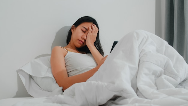 A mulher asiática nova que usa o smartphone que verifica os meios sociais que sentem o sorriso feliz ao encontrar-se na cama após acorda na manhã, sorriso latino-americano atrativo bonito da senhora relaxa no quarto em casa.