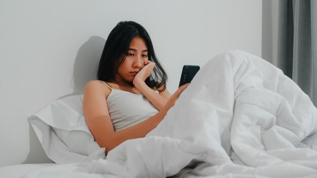 A mulher asiática nova que usa o smartphone que verifica os meios sociais que sentem o sorriso feliz ao encontrar-se na cama após acorda na manhã, sorriso latino-americano atrativo bonito da senhora relaxa no quarto em casa.