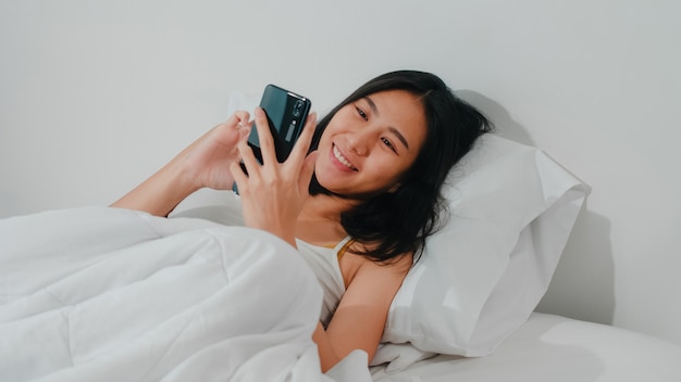 A mulher asiática nova que usa o smartphone que verifica os meios sociais que sentem o sorriso feliz ao encontrar-se na cama após acorda na manhã, sorriso latino-americano atrativo bonito da senhora relaxa no quarto em casa.