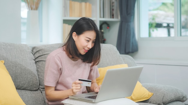 A mulher asiática que usa o comércio eletrônico da compra do portátil e do cartão de crédito, fêmea relaxa sentindo a compra online feliz que senta-se no sofá na sala de visitas em casa. As mulheres do estilo de vida relaxam em casa o conceito.