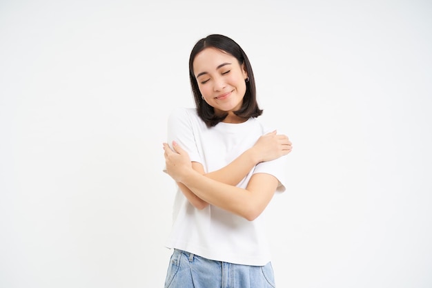 A mulher asiática sorridente e feliz do amor-próprio se abraça sobre o fundo branco