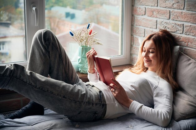 A mulher está deitada na cama do peitoril da janela e lendo o livro