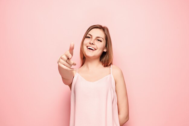 A mulher feliz aponta e quer você, retrato de meio comprimento closeup na parede rosa