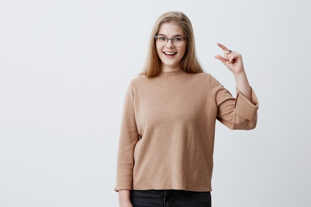 A mulher loura bonita contente nos monóculos mostra algo pequeno com mãos, veste a camisola marrom, isolada contra o fundo cinzento do estúdio. Bela jovem fêmea demonstra o tamanho de algo