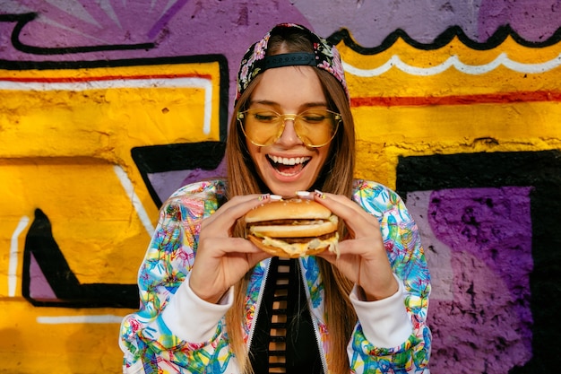 A mulher nova bonita feliz, sorrindo extensamente, prende o hamburguer saboroso em duas mãos.