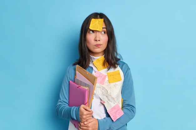 A mulher pensativa de Asain tem notas adesivas sobre roupas e testa pensativa trabalha duro durante o prazo de entrega de pastas com documentos.