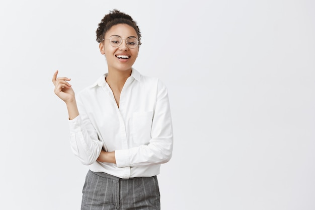 A mulher pode lidar com qualquer cliente. Mulher africana atraente, confiante e feminina, com cabelos cacheados, gesticulando com a mão levantada, sorrindo com expressão autoconfiante, vestindo terno e óculos