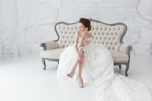 A noiva no vestido longo que senta-se no sofá dentro no interior branco do estúdio gosta em casa.