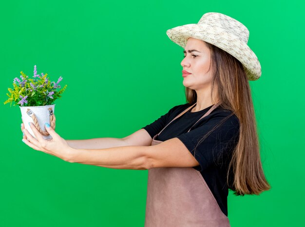 A pensar na bela jardineira de uniforme usando chapéu de jardinagem, segurando uma flor em um vaso de flores ao lado isolado no fundo verde