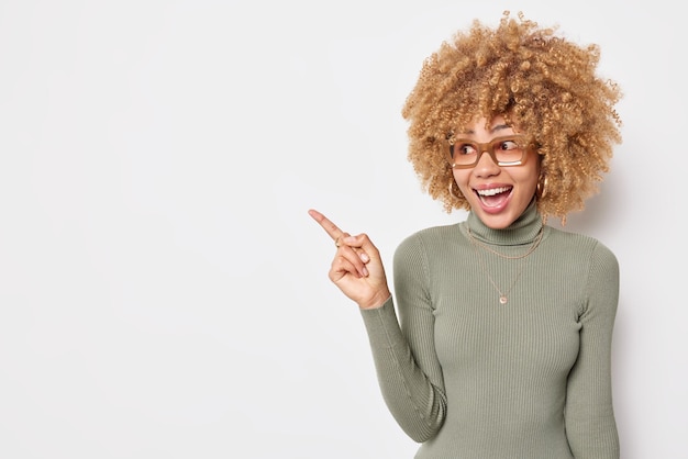 A promotora feliz e surpresa aponta para um espaço em branco e mostra algo incrível. Demonstra uma simulação de lugar que usa óculos e gola olímpica isolada sobre fundo branco.