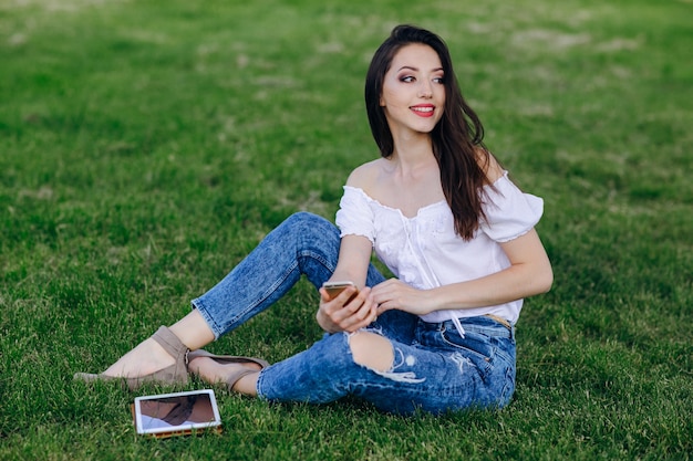 A rapariga sentado em um parque com uma tabuleta na grama e um celular na mão