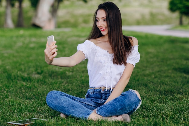 A rapariga sentado em um parque fazendo uma foto auto ao sorrir