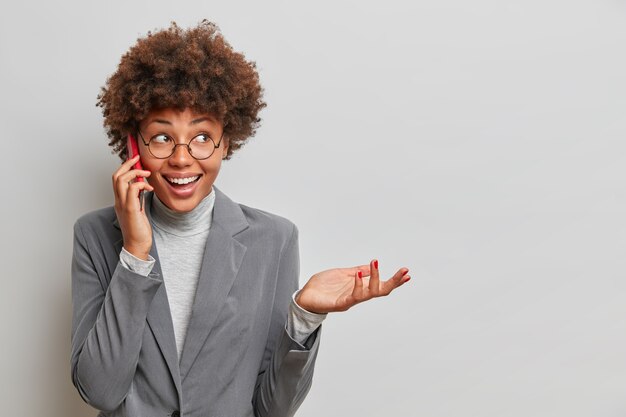 A secretária afro-americana feliz discute questões de trabalho com colega de trabalho via smartphone, recebe ligação do gerente, levanta a mão e ri alegremente, informa sobre estágio de trabalho