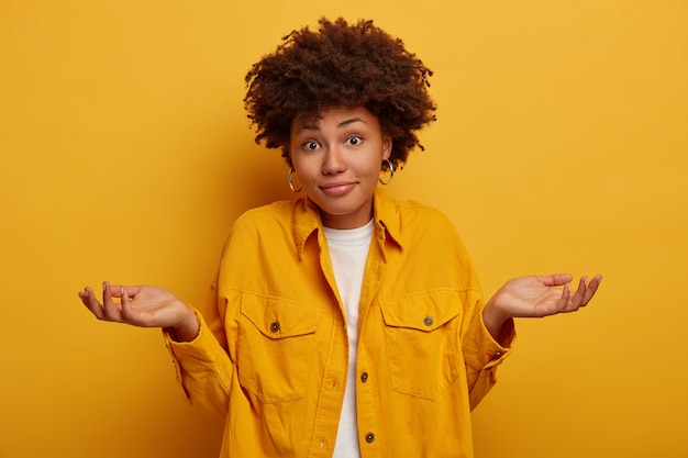 A senhora africana encolhe os ombros e expressa incerteza, toma decisões, veste roupas da moda, estende as palmas das mãos para os lados isoladas na parede amarela.