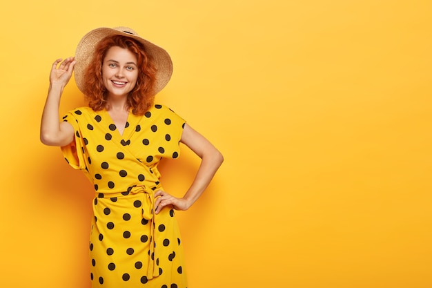 A senhora alegre do verão segura uma mão na cintura e a outra no chapéu de palha, usa um vestido de bolinhas amarelo vívido, tem uma aparência feliz, corpo esguio, modelos internos, copie o espaço certo para sua promoção. beleza e feminilidade