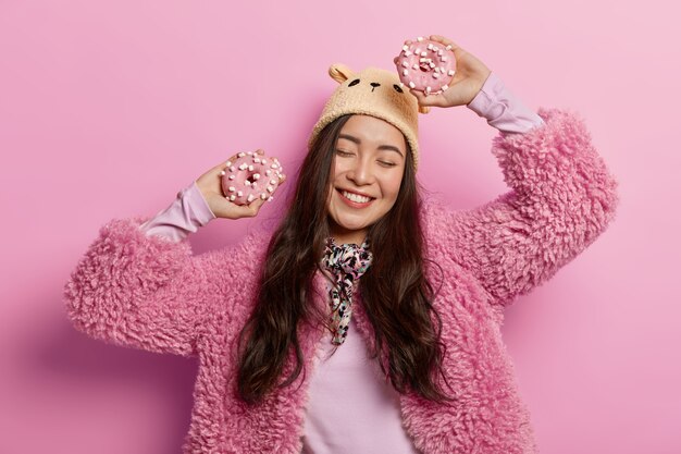 A senhora despreocupada dança alegremente com dois donuts doces, se diverte dentro de casa, usa casaco rosa, chapéu marrom