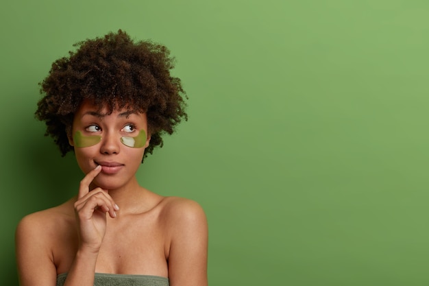 A senhora pensativa cuida da pele e do corpo, aplica tapa-olhos após o banho, mantém os dedos perto dos lábios, faz tratamento de rejuvenescimento, cosmetologia anti-envelhecimento, estande interior