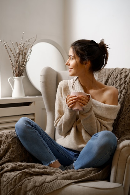 A senhora sorridente em roupas da moda inteligente está sentada na poltrona com uma xícara de chá.