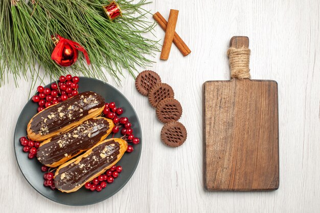 A vista de cima éclairs de chocolate e groselha na placa cinza. Cookies cruzaram folhas de canela e pinheiro com brinquedos de Natal e uma tábua de cortar na mesa de madeira branca