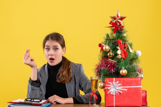 A vista de frente surpreendeu a garota sentada na mesa mostrando um gesto de dinheiro, árvore de natal e coquetel de presentes