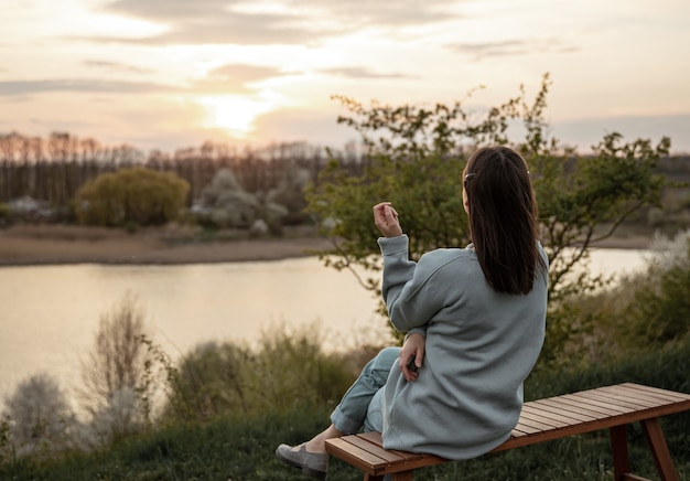 A vista de trás da menina olha para o pôr do sol, sentado em um banco.