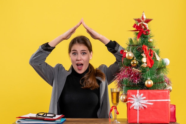 A vista frontal surpreendeu a garota sentada na mesa de mãos dadas como uma casa no telhado perto da árvore de natal e coquetel de presentes