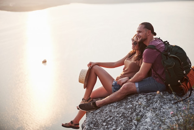 Foto grátis abraçando o casal com mochila sentado no topo da montanha de pedra, apreciando a vista costeia um rio ou lago.