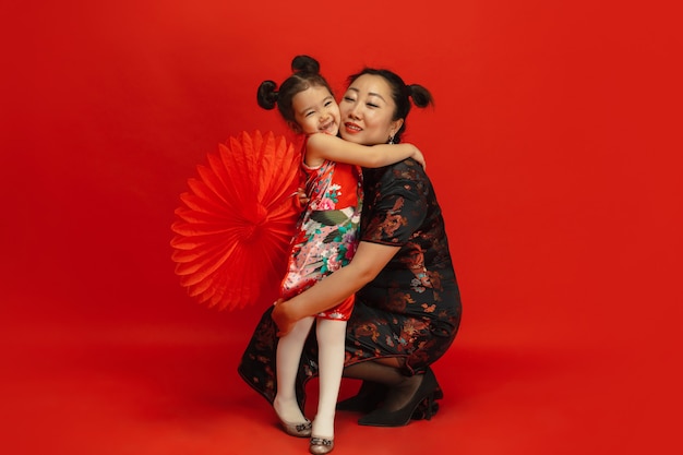 Abraçando, sorrindo feliz. Feliz ano novo chinês 2020. Retrato de mãe e filha asiático isolado em um fundo vermelho em roupas tradicionais. Celebração, emoções humanas, feriados. Copyspace.