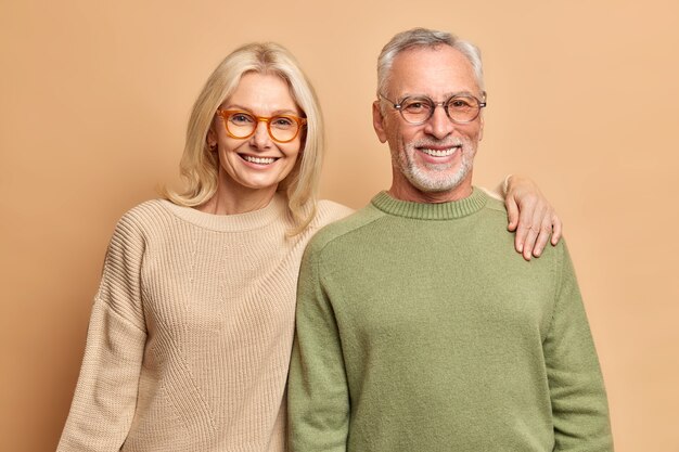 Abraço de casal maduro sorridente olha com alegria para a pose da câmera para o retrato de família. Crianças felizes vieram visitá-los usando óculos transparentes. Jumpers casuais isolados sobre a parede marrom