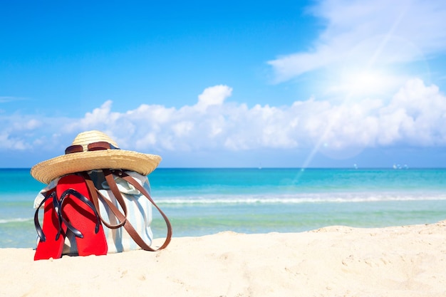Acessórios de praia na areia para o conceito de férias de verão. bolsa, chapéu de palha com óculos escuros e chinelos vermelhos. areia branca com incrível oceano e céu azul ao fundo. espaço livre para seu texto. Foto Premium