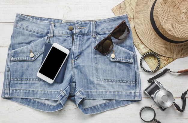 Acessórios de vista superior para viajar com conceito de roupa feminina. Telefone móvel, chapéu, mapa, câmera, colar, calças e óculos de sol na mesa de madeira branca.