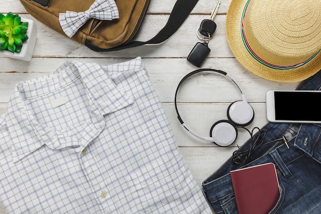 Acessórios de vista superior para viajar com o conceito de vestuário do homem. Camisa branca, jean, telefone móvel, ouvindo música por fone de ouvido em fundo de madeira. Bolsa, passaporte, chave, óculos de sol e chapéu na mesa de madeira.