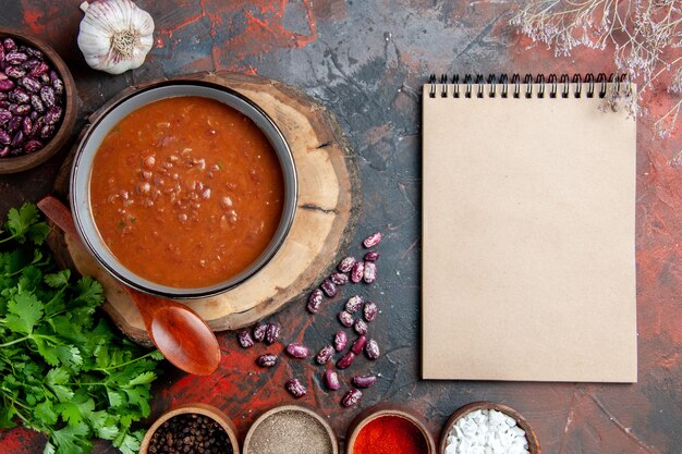 Acima, vista da clássica sopa de tomate em uma tigela azul colher na bandeja de madeira alho sal um monte de verde e caderno na mesa de cores misturadas