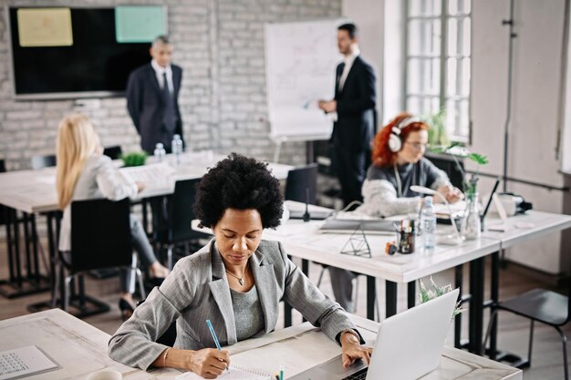 Acima vista da empresária afro-americana usando computador e escrevendo notas no papel no escritório