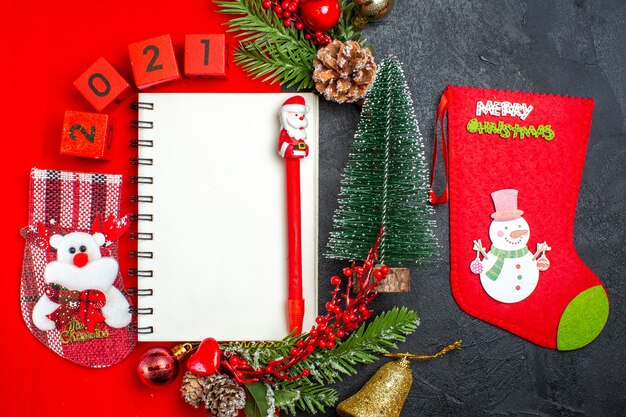 Acima, vista de acessórios de decoração de caderno espiral ramos de abeto xsmas números de meias em um guardanapo vermelho e árvore de Natal em fundo escuro