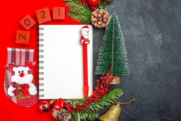 Acima, vista de acessórios de decoração de caderno espiral ramos de abeto xsmas números de meias em um guardanapo vermelho e árvore de Natal em fundo escuro