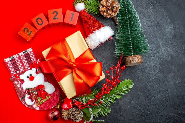 Acima, vista de belos acessórios de decoração de presentes ramos de abeto xsmas números de meias em um guardanapo vermelho e árvore de Natal chapéu de Papai Noel em fundo escuro