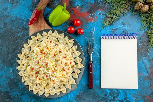 Acima, vista de macarrão farfalle italiano cru com legumes em uma placa preta na tábua de madeira e um garfo ao lado do caderno espiral de tomates de pimentão sobre fundo azul