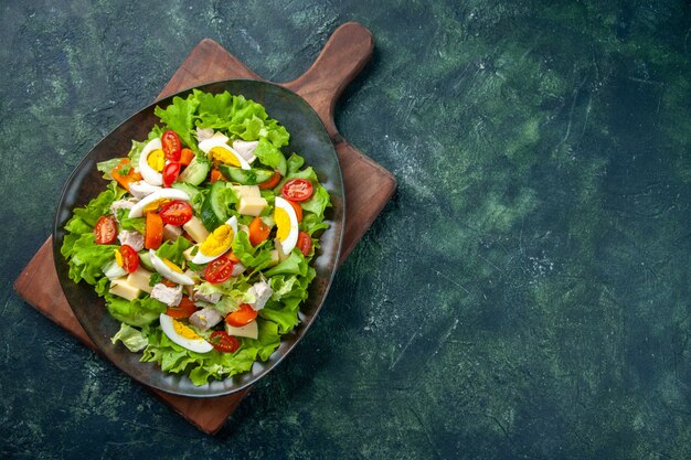 Acima, vista de uma deliciosa salada com muitos ingredientes frescos no lado direito na tábua de madeira na mesa de cores pretas e verdes