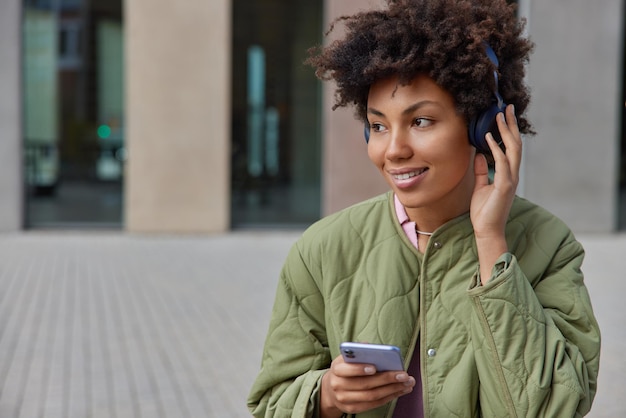 Adolescente alegre de cabelos cacheados recria nas horas vagas satisfeita com som de qualidade em fones de ouvido, usa aplicativo de celular vestida com roupas casuais