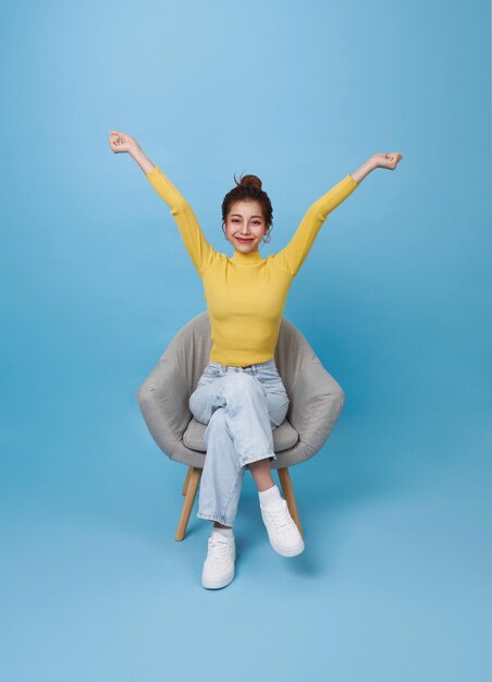 Adolescente asiática atraente sentada em uma cadeira e relaxar a mão para cima isolada em fundo azul