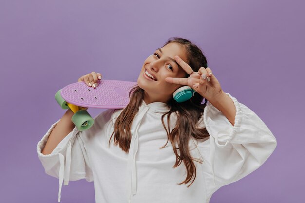 Adolescente bronzeado mostra sinal de paz e ouve música em fones de ouvido azuis Jovem feliz segura longboard e sorri em fundo roxo