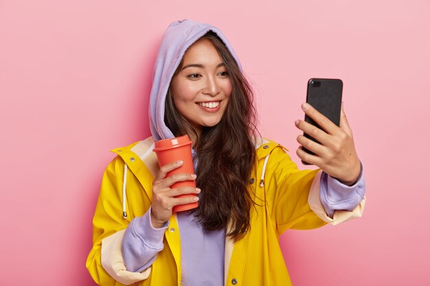 Adolescente com aparência específica tira uma selfie, caminha ao ar livre durante o dia de outono, usa capa de chuva protetora e bebe café no frasco