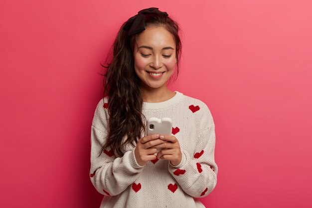 Adolescente com sorriso gentil, digita mensagens no celular moderno, conversa online, lê postagens nas redes sociais, viciada em tecnologia