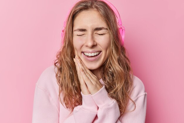 Adolescente de cabelos compridos muito feliz mantém as mãos pressionadas ri alegremente fecha os olhos ouve música via fones de ouvido estéreo sem fio usa moletom casual isolado sobre fundo rosa