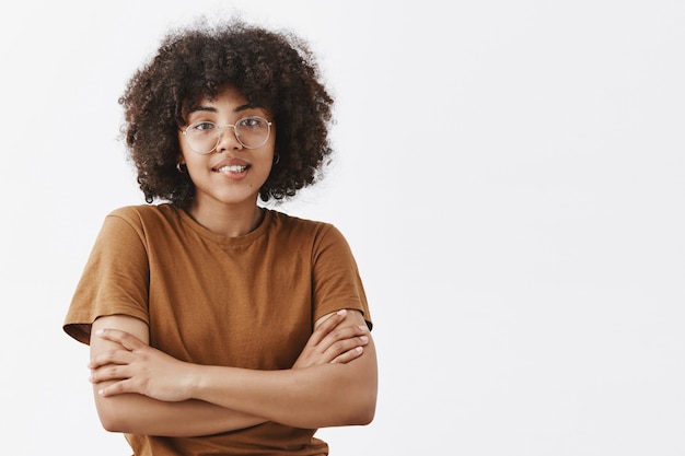 adolescente de pele escura usando óculos transparentes e uma camiseta marrom da moda, sentindo frio cruzando as mãos no peito e sorrindo com uma expressão amigável e educada