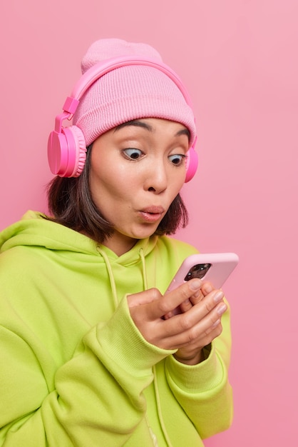 Foto grátis adolescente espantada com o olhar surpreso com smartphone estressado com a leitura de más notícias recebe recibo de conta usando chapéu e moletom poses contra a parede rosa